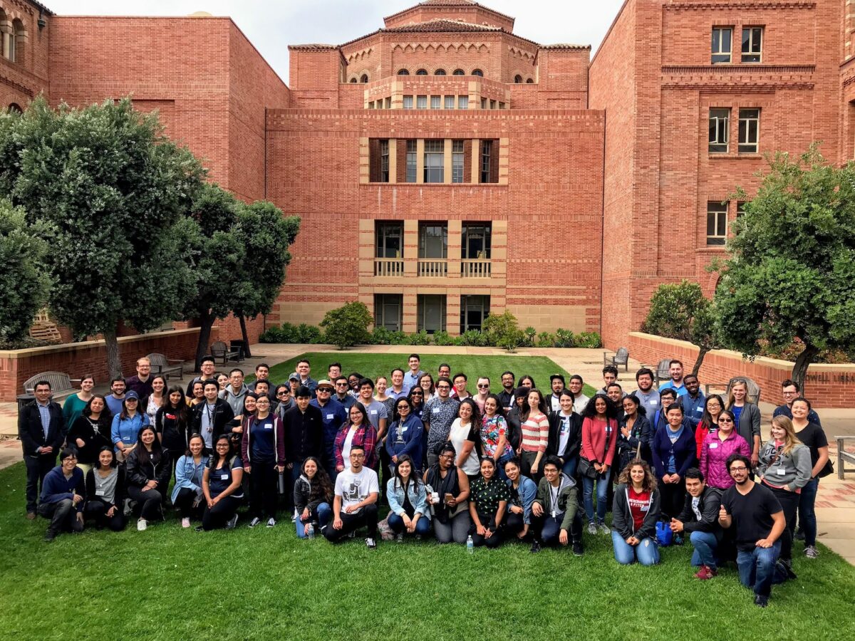 An image of a group of physical science student, faculty, and staff sitting on the lawn in front of the UCLA campus