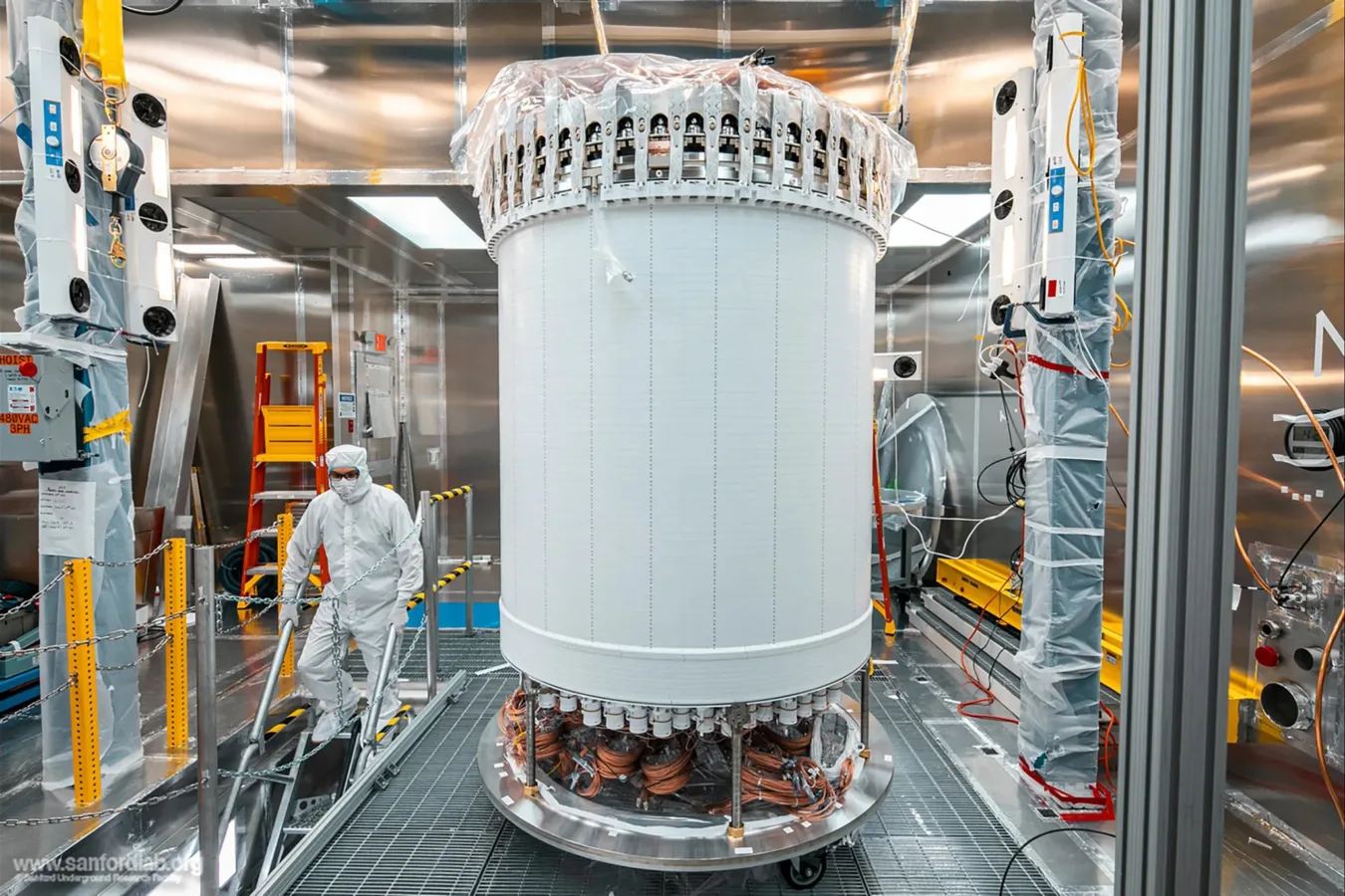 A researcher in protective gear works near a tall, cylindrical scientific instrument with visible wiring. The walls surrounding it are reflective metal.