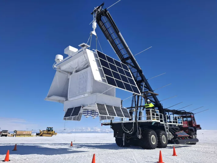 Photo of a crane lifting the GAPS instrument. Other heavy machinery is in the background, the ground is hard-packed snow/ice, and the sky is cloudless and deep-blue.