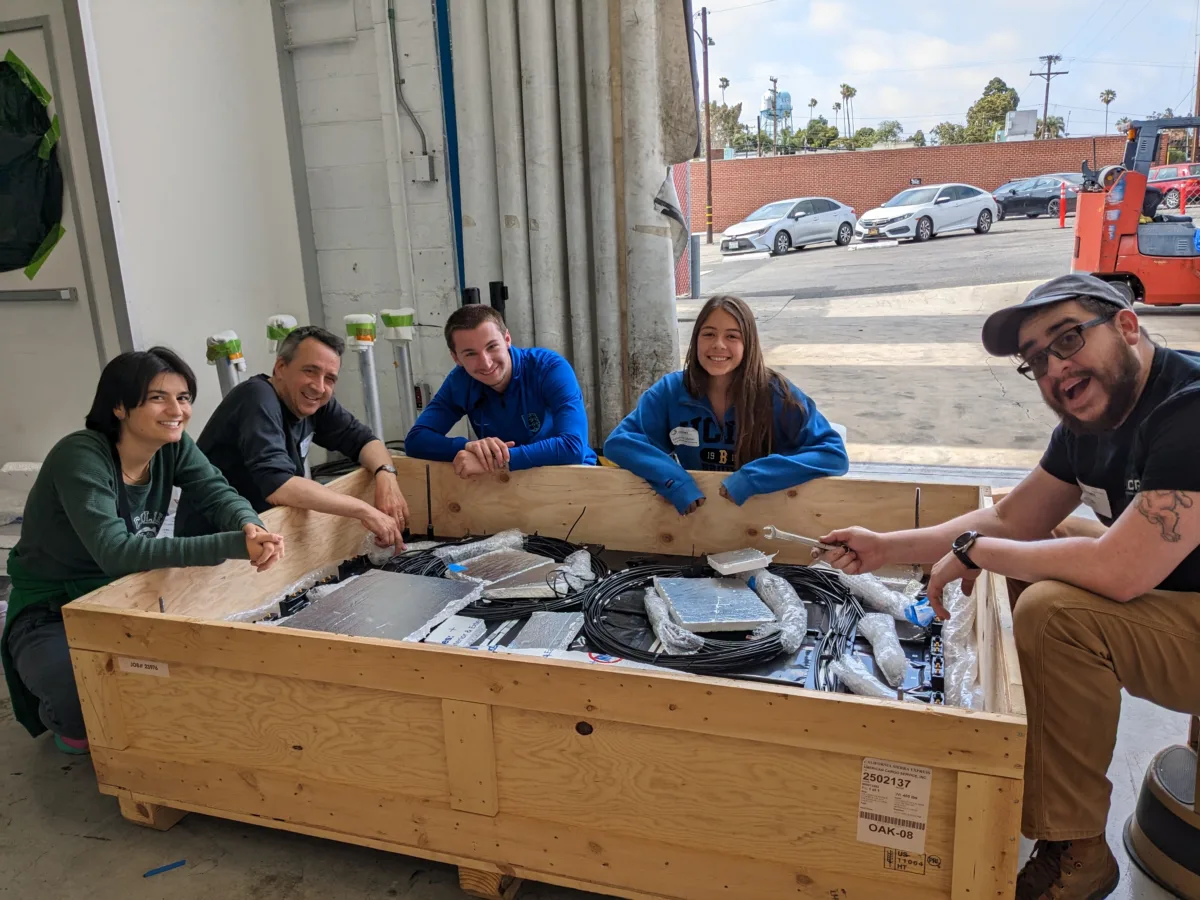 Five people sit surrounding a large wooden crate. The crate contains scientific equipment related to the GAPS instrument.