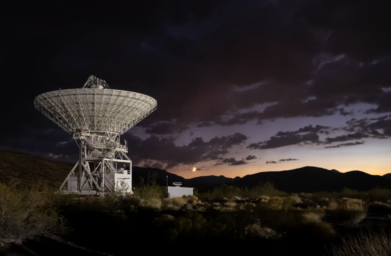 Deep Space Station 15 (DSS-15), one of the 112-foot (34-meter) antennas at the Goldstone Deep Space Communications Complex near Barstow, California, is seen at sunset in September 2025. The crescent Moon hangs just above the horizon. Goldstone is part of NASA’s Deep Space Network (DSN), which operates three complexes around the globe that support communications with dozens of deep space missions.